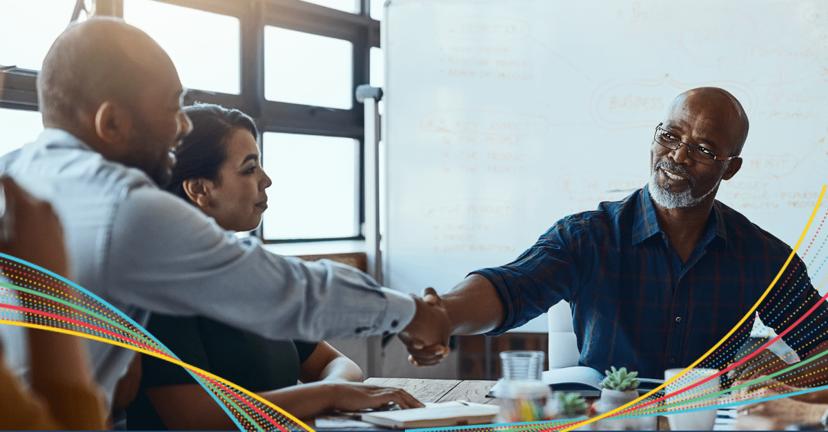 3 team members at a table while two handshake in an office